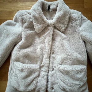 Beige teddyjacka från H&M - Mysig beige teddyjacka från H&M med knappar framtill och stora fickor. Perfekt för kyliga dagar när du vill hålla dig varm och stilren. Jackan har en klassisk krage och långa ärmar.