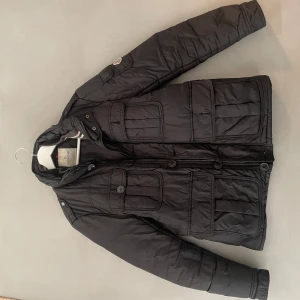 Svart quiltad jacka från Moncler - Snygg svart quiltad jacka från Moncler med långa ärmar och flera praktiska fickor. Jackan har både dragkedja och knappar framtill samt ett broderat Moncler-märke på ärmen. Perfekt för kyligare dagar. Skick 6/10 det saknas några knappar vid fickorna men går att sy på och avvisen är nästan av jag har kvitto köpt av trendsellers 