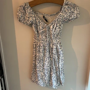 Blommig klänning från BikBok - Söt blommig klänning från BikBok i vitt och blått. Klänningen har korta puffärmar och en smockad överdel som ger en fin passform. Perfekt för sommarens varma dagar!