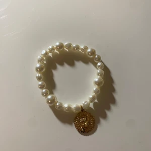 Pärlarmband med guldberlock - Elegant pärlarmband med vita pärlor och en rund guldberlock. Perfekt för att ge en touch av klass till din outfit. Berlocken har en detaljerad design som ger en extra lyxig känsla. Priset går att diskutera💗