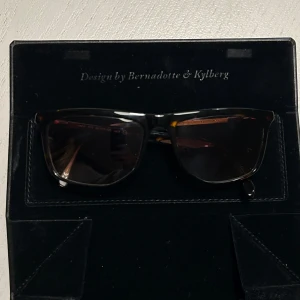 Solglasögon från Bernadotte & Kylberg - Snygga solglasögon designade av Bernadotte & Kylberg. De har en klassisk rektangulär form med mörka linser och en elegant båge. Perfekta för att ge en stilren touch till din look.