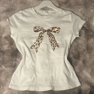 Vit t-shirt med leopardmönstrad rosett från Stradivariu - Säljer en söt vit t-shirt från Stradivarius med en stor leopardmönstrad rosett på framsidan. T-shirten har en tight passform och korta ärmar. Perfekt för att ge en touch av stil till din vardagslook! Nästan ny skick, bara använd några gånger.