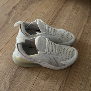 Nike Air Max 270 - Säljer ett par vita Nike sneakers med en modern design och perforerade detaljer för extra ventilation. Skorna har en synlig luftkudde i hälen för komfort och stil. Perfekta för både träning och vardag.