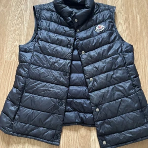 Svart dunväst från Moncler - Snygg svart dunväst från Moncler med knappar och ett broderat märke på bröstet. Perfekt för kyliga dagar när du vill hålla stilen. Västen har en klassisk design med quiltade paneler.