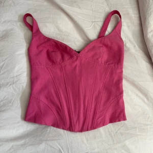 Rosa korsettopp - Säljer en snygg rosa korsettopp med smala axelband och dragkedja i sidan. Toppen har en smockad detalj på baksidan för extra stretch och komfort. Perfekt för att ge en stilren och feminin look. Axelbanden är kortade!!