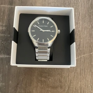 Silverfärgad klocka från Calvin Klein - Elegant klocka från Calvin Klein med en stilren design. Klockan har en silverfärgad boett och armband, samt en mörk urtavla med minimalistiska markeringar. Perfekt för en tidlös look. Oanvänd
