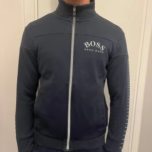 Mörkblå kofta med dragkedja från Hugo Boss - Snygg mörkblå kofta från Hugo Boss med dragkedja och hög krage. Tröjan har vita detaljer på ärmarna och logotypen tryckt på bröstet. Priset kan diskuteras vid snabb affär 