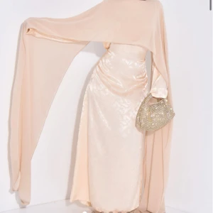 Champagnefärgad långklänning med cape - Elegant champagnefärgad långklänning med en dramatisk cape som ger en flytande effekt. Klänningen har en omlott-design och är tillverkad i ett glansigt material som ger en lyxig känsla. Perfekt för speciella tillfällen. Aldrig andvänd!