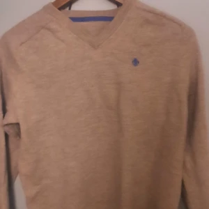 Beige tröja med v-ringning - Säljer en stilren beige tröja med v-ringning och ett litet blått broderat emblem på bröstet. Perfekt för en avslappnad men ändå snygg look. Tröjan har långa ärmar och är gjord i ett mjukt material.