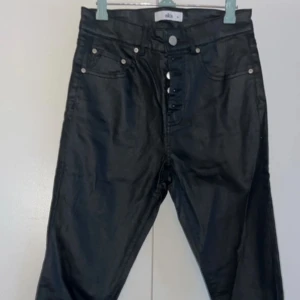 Svarta byxor med knappar - Snygga svarta byxor med knappar framtill. De har en stilren design och passar perfekt för en modern look. Byxorna har en normal passform och är tillverkade i ett glansigt material som ger en extra touch.