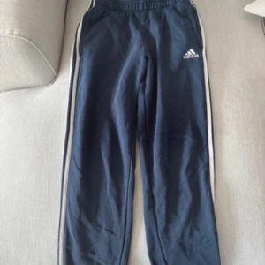 Blå mjukisbyxor från Adidas - blåa träningsbyxor från Adidas med vita ränder längs sidorna. Byxorna har en elastisk midja och är perfekta mysbyxor. Adidas-loggan är tryckt på ena benet.