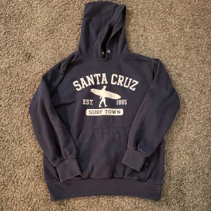 Mörkblå hoodie med Santa Cruz-tryck - Säljer en mörkblå hoodie från Santa Cruz med ett stort tryck på framsidan. Hoodien har en klassisk känguruficka och en bekväm huva.