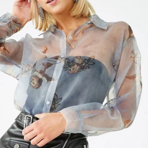Transparent blus med änglamotiv - Säljer en unik transparent blus med ett vackert änglamotiv och blommönster. Blusen har långa ärmar och knappar framtill. Perfekt för att ge en konstnärlig touch till din outfit. Det saknas en knapp på toppen.  
