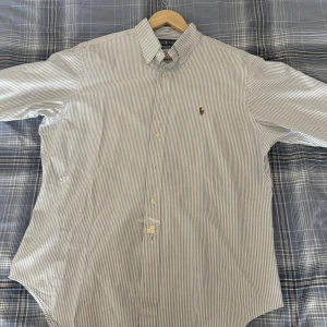 Vit och blå randig skjorta från Ralph Lauren - Snygg vit och blå randig skjorta från Ralph Lauren med klassisk krage och broderad logga på bröstet. Skjortan är helt oanvänd och säljs då den är för stor. För fler bilder, hör av dig.