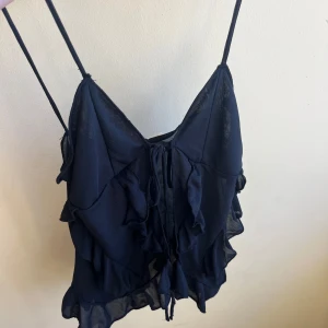 Svart linnetopp från Pull&Bear - Snygg svart linnetopp från Pull&Bear med smala axelband och volangdetaljer. Toppen har en knytning framtill och är perfekt för en stilren look. Aldrig använd, nyskick utan prislapp.