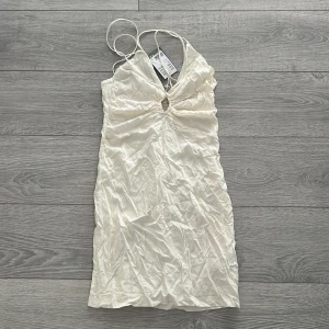 Vit  klänning - Säljer en elegant vit klänning med en v-ringad design och en dekorativ detalj framtill. Klänningen har en kort längd och är perfekt för sommarens festligheter. Materialet är mjukt och följsamt, vilket ger en bekväm passform.