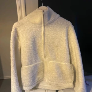 Vit teddyjacka från 157 - Mysig vit teddyjacka från 157 med dragkedja framtill och två stora fickor. Perfekt för kyliga dagar med sin fluffiga och varma design. Jackan har en hög krage och långa ärmar för extra komfort. Små tecken på användning 