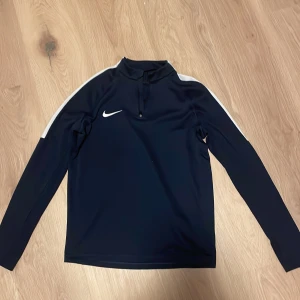Svart långärmad tröja från Nike - Säljer en svart långärmad tröja från Nike med vit rand längs ärmarna. Tröjan har en halv dragkedja vid halsen och är tillverkad i Dri-FIT material för optimal komfort. Perfekt för träning eller vardagsbruk.