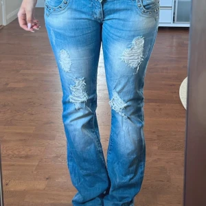 Blå bootcut jeans med slitningar - Snygga blå bootcut jeans från Adoro med slitningar på framsidan och dekorativa nitar på fickorna. Jeansen har en låg midja. Dom är nästan helt oanvända och jag köpte dem för 1400kr.  Står inte storlek men jag att skulle säga M