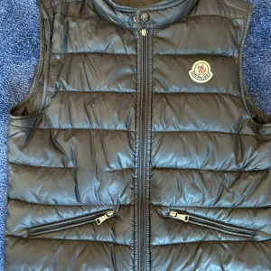 Svart dunväst från Moncler storlek 152 - Snygg svart dunväst från Moncler med dragkedja och två fickor med dragkedjor. Perfekt för kyligare dagar och ger en stilren look. Moncler-loggan pryder bröstet för en exklusiv touch.