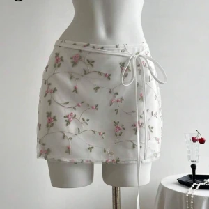 Vit blommig kjol med knytband - Charmig vit kortkjol med blommigt broderi i rosa och grönt. Kjolen har ett knytband i midjan för en justerbar storlek. Helt oanvänd. Storlek m, passar även smal. 