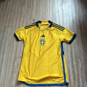 Gul fotbollströja från Adidas - Säljer en gul fotbollströja från Adidas med Sveriges landslagsemblem på bröstet. Tröjan har korta ärmar och blå detaljer, inklusive ränder på axlarna och runt ärmsluten. Perfekt för fotbollsfans som vill visa sitt stöd för landslaget!ny pris 300kr priset får även att diskuteras 