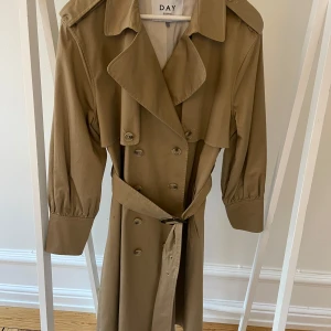 Beige trenchcoat från Day Birger et Mikkelsen - Snygg beige klassisk trenchcoat från Day Birger et Mikkelsen med klassisk design. Jackan har knappar framtill och ett bälte i midjan för en elegant look. Perfekt för vår och höst.