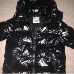 Svart pufferjacka från Moncler - Snygg svart pufferjacka från Moncler med glansig finish och dragkedja framtill. Jackan har en huva och praktiska fickor med dragkedjor. Perfekt för kyliga dagar.