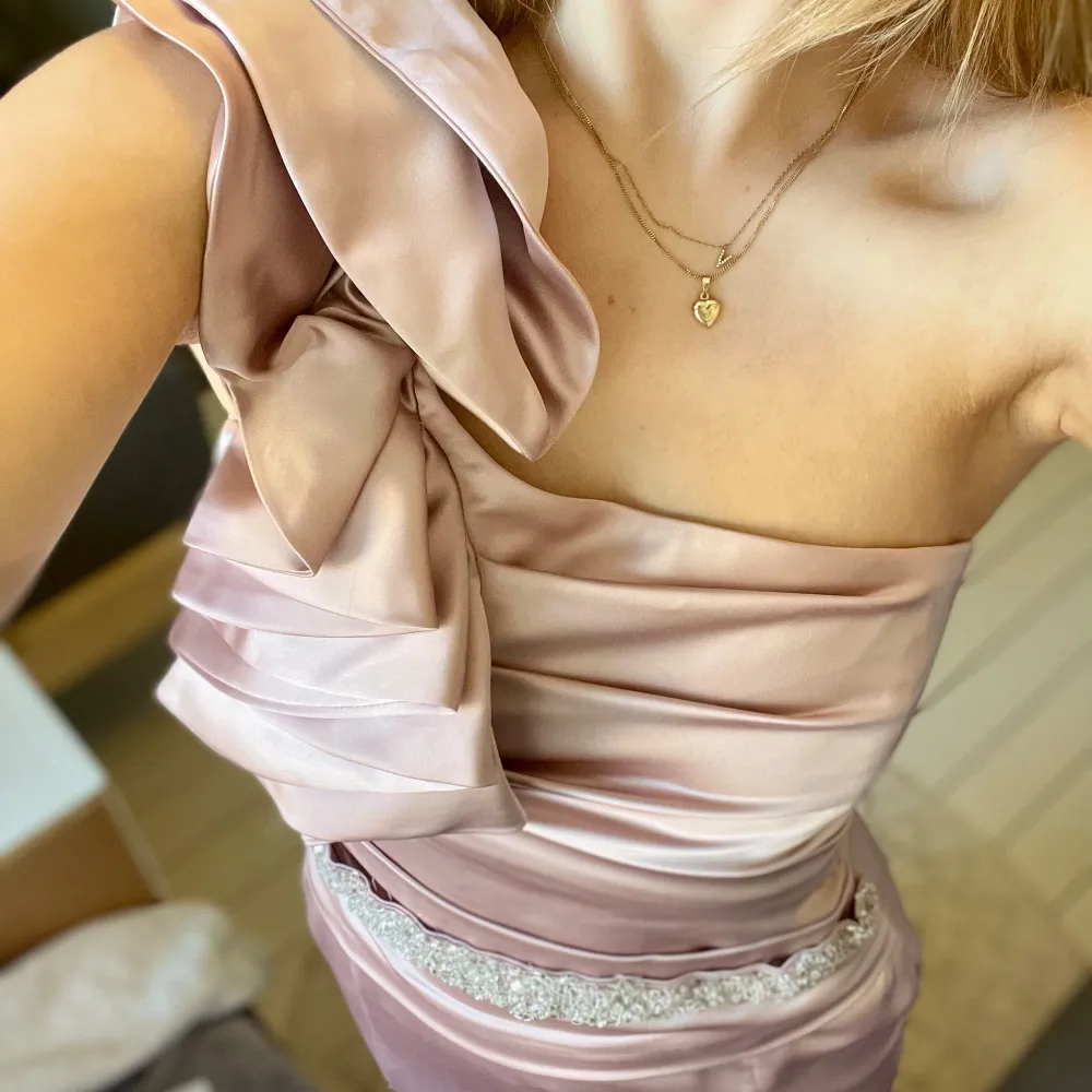 Elegant champagnefärgad balklänning i satin med ”One-shoulder” design. Klänningen har en glittrigt band(går att ta av). Se sista bild för en liten missfärgning.. Mekot.