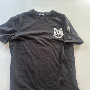 Svart t-shirt från Moncler - Säljer en stilren svart t-shirt från Moncler med ett stort broderat M-logotyp på bröstet och Moncler-märke på ärmen. Perfekt för en casual look. T-shirten är kortärmad och gjord i mjukt bomullsmaterial.