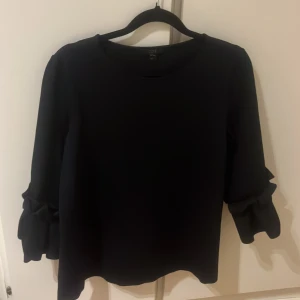 Svart blus från COS - Säljer en elegant svart blus från COS med unika volangdetaljer på ärmarna. Perfekt för en stilren look. Blusen har trekvartsärmar.