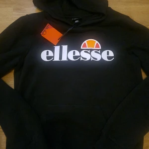 Svart hoodie från Ellesse - Säljer en svart hoodie och byxor från Ellesse  med deras ikoniska logga i vitt och orange på bröstet. Perfekt för en avslappnad stil med en klassisk look. Hoodien har långa ärmar och en bekväm passform.......  sköna snygga svarta byxor till    