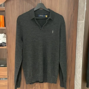 Grå långärmad piké Ralph Lauren - Säljer en stilren mörkgrå tröja från Ralph Lauren. Tröjan har en klassisk krage och knappar vid halsen. Den är långärmad och har en diskret logotyp broderad på bröstet. Perfekt för en elegant och avslappnad look. Sparsamt använd och håller storlek bra. Hör av er vid minsta intresse!🤩