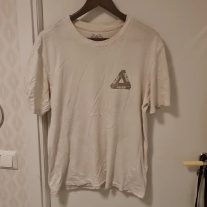 Vit t-shirt från Palace - Säljer en vit t-shirt från Palace med deras ikoniska triangellogga tryckt på både fram- och baksidan. T-shirten har en normal passform och är kortärmad, perfekt för en avslappnad stil.