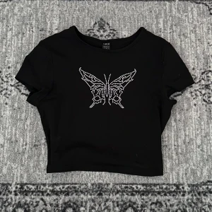 Svart topp med fjärilsdetalj - Snygg svart croppad topp med en glittrig fjäril i strass på framsidan. ✨