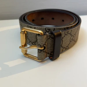 Mönstrat bälte från Gucci - Snyggt bälte från Gucci med ett klassiskt GG-mönster och en cool tigerdetalj. Bältet har ett guldfärgat spänne med märkets logotyp. Perfekt för att ge din outfit en lyxig touch. Köpt på garmsmarket för ett år sedan ungefär så antar äktheten. 