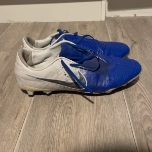 Blå och vita fotbollsskor från Nike - Säljer ett par Nike fotbollsskor i blått och vitt med snörning. Skorna har en rund tå och är designade för att ge bra grepp på planen. Perfekta för fotbollsträning och matcher.