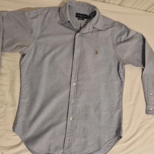 Blå skjorta från Ralph Lauren - Snygg ljusblå skjorta från Ralph Lauren med klassisk krage och broderad logga på bröstet. Skjortan har långa ärmar och knappar framtill. Perfekt för en stilren look. Inga defekter. Tveka inte att höra av dig, priset är inte hugget i sten.👌🔥