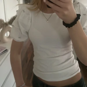 Vit kortärmad topp med puffärmar - Säljer en vit kortärmad topp med puffiga ärmar och dekorativa detaljer som är köpt på NewYorker Toppen har en tight passform och är perfekt för en stilren look. Den är gjord i ett mjukt material som känns skönt mot huden.