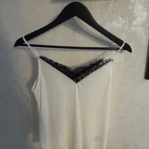 Vit linne med spetsdetaljer - Säljer ett stilrent vitt linne med svarta spetsdetaljer vid halsringningen. Linnet har smala axelband och en elegant v-ringning. Perfekt för en chic och enkel look.