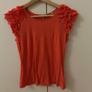 Korallfärgad topp med volangärmar - Säljer en snygg korallfärgad toppmed unika volangdetaljer på ärmarna. Perfekt för att liva upp garderoben med lite färg och stil. Toppen har en rund halsringning och är tillverkad i ett mjukt material.