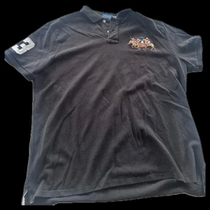 Svart pikétröja från Polo Ralph Lauren - Säljer en svart pikétröja från Polo Ralph Lauren med broderat polospelmotiv på bröstet och siffran 3 på ärmen. Tröjan har en klassisk krage och knappar framtill. Perfekt för en stilren look.