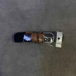 Blått flätat bälte med bruna detaljer - Snyggt svart flätat bälte med bruna läderdetaljer och silverfärgat spänne. Perfekt för att ge en stilren touch till din outfit. Bältet är förpackat i plast och har en klassisk design.