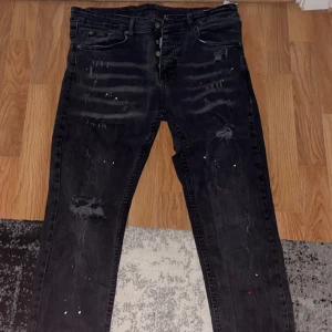 Svarta jeans från Dsquared2 - Snygga svarta jeans från Dsquared2 med slitna detaljer och knappgylf. Jeansen har en cool, distressed look med små färgstänk och en röd logotyp på baksidan. Perfekta för en trendig stil. Jeansen är slimfit och är använda några gånger. Jeansen är köpta på hemsidan disquared.