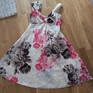 Blommig klänning med rosa och grå rosor - Säljer en vacker klänning med blommigt mönster i rosa och grått. Klänningen har en ärmlös design och en elegant passform. Perfekt för sommarens festligheter!