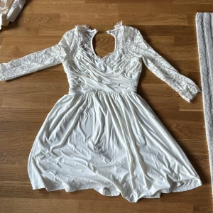 Vit spetsklänning från Chiara Porthi - Säljer en elegant vit klänning från Chiara Porthi med vackra spetsdetaljer och trekvartsärmar. Klänningen har en omlott-design vid bysten och en kort kjol som ger en romantisk look. Perfekt för speciella tillfällen!