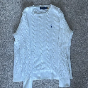 Vit stickad tröja från Ralph Lauren - Säljer en klassisk vit stickad tröja från Ralph Lauren med kabelstickat mönster och den ikoniska loggan broderad i blått på bröstet. Tröjan har långa ärmar och en rund halsringning. Perfekt för en stilren look!