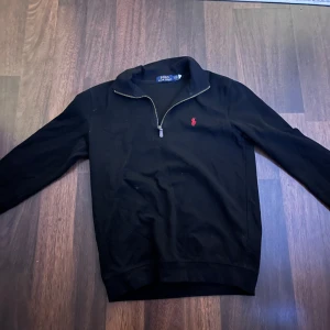 Svart tröja med dragkedja från Polo Ralph Lauren - Säljer en stilren svart tröja från Polo Ralph Lauren med en halv dragkedja och röd logga på bröstet. Perfekt för en avslappnad men snygg look. Tröjan har långa ärmar och är tillverkad i ett mjukt material. 