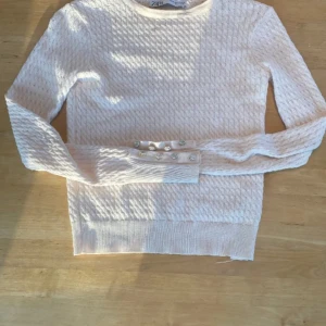 Ljusrosa stickad tröja från Zara - Säljer en stilren ljusrosa stickad tröja från Zara med långa ärmar och dekorativa pärlknappar på ärmsluten. Perfekt för en elegant look. Tröjan har en fin textur och ribbade kanter.