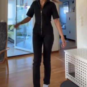 Svart/grå jeans jumpsuit  - Snygg svart jumpsuit med korta ärmar och dragkedja framtill. Den har en figurnära passform och är perfekt för en stilren look. Jumpsuiten är enkel att styla och passar till många tillfällen! Den är från mango, har enbart en liten missfärgad fläck där de åkt bort lite svårt färg på benet men inget som märks. 